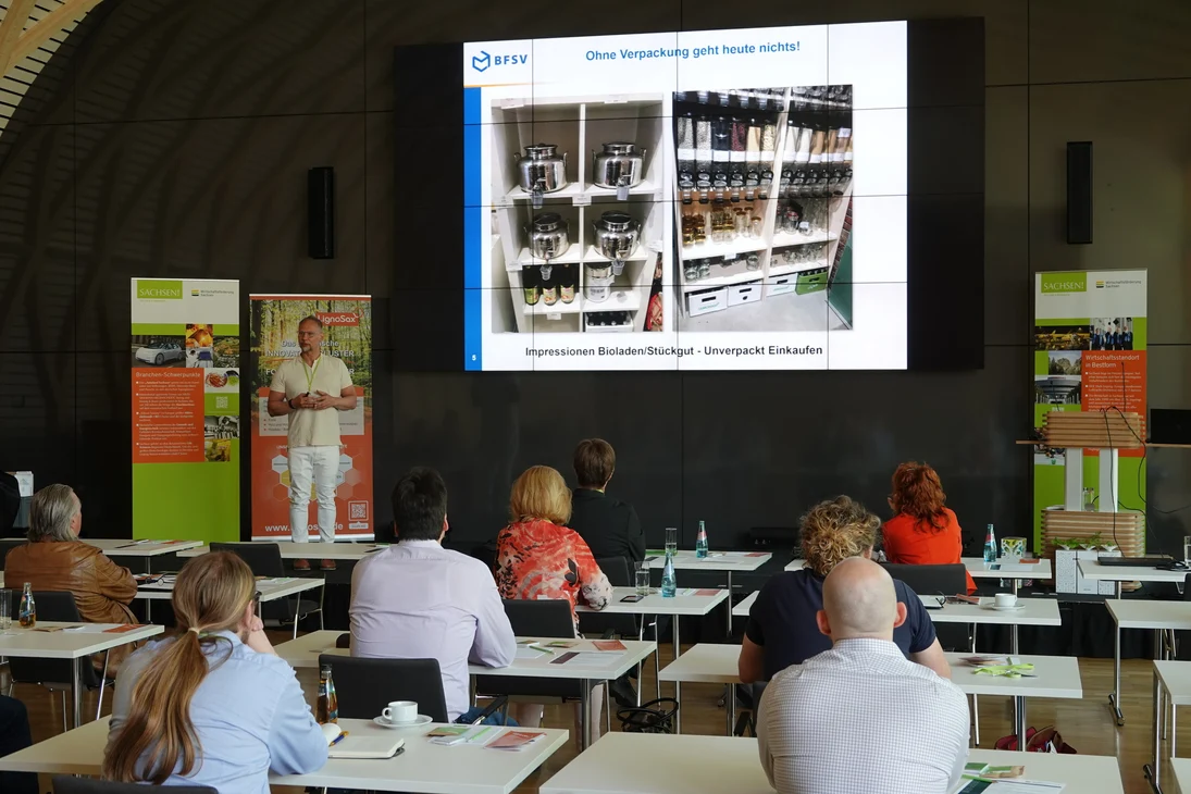 Ein Mann steht vor einer Leinwand mit Präsentationsfolie. Mehrere Personen hören ihm zu