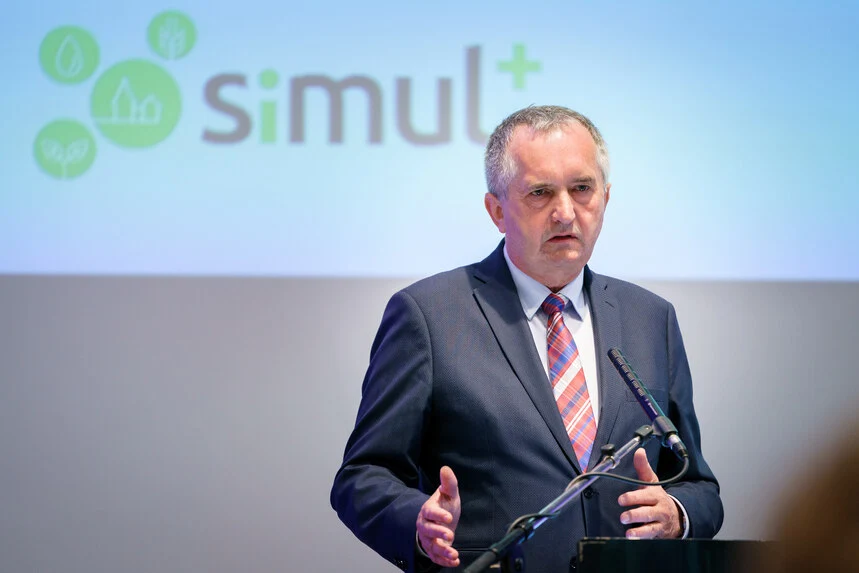 Staatsminister Schmidt steht an einem Pult und spricht in ein Mikrofon. Im Hintergrund ist das grüne simul+ Logo zu sehen.