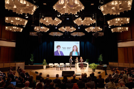 Blick in den Saal mit vielen Zuschauern. Auf der Bühne in der Mitte des Raumes stehen zwei Frauen und ein Mann an einer Stehbrücke und diskutieren.
