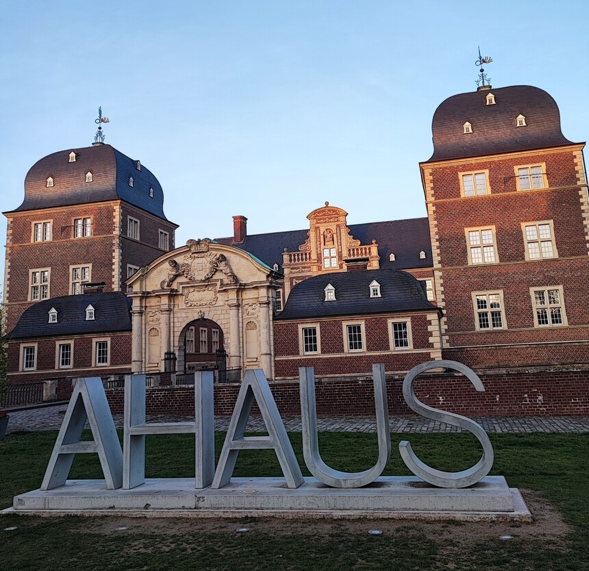 Vor einem Schloss ist eine grüße Wiese mit dem Schriftzug Ahaus in sehr großen Buchstaben zu sehen.