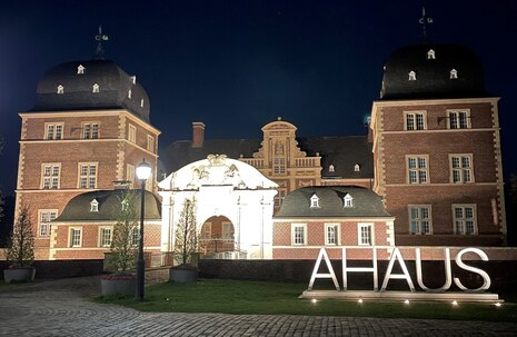 Blick bei Nacht auf ein Schloss welches beleuchtet ist und der Schriftzug Ahaus davor aufgestellt wurde.