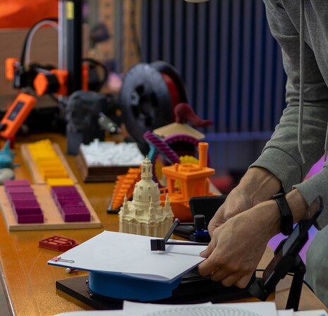 Auf einem Tisch liegen Materialien zum Basteln und Handwerken sowie ein Modell der Frauenkirche Dresden. Auf der rechten Seite ragen zwei Hände eines Mannes ins Bild, der mit den Materialien arbeitet.