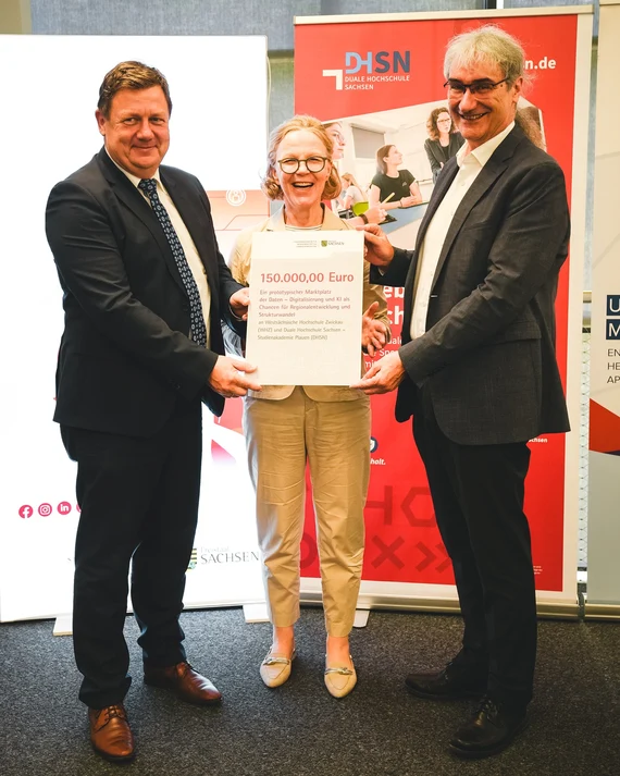 Ministerin Regina Kraushaar und zwei Männer stehen vor Bannern und halten einen Fördermittelbescheid in der Hand.