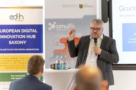 Ein Mann steht vor einem Tisch und spricht in ein Mikrofon und hält eine Chipkarte in der Hand. Im Hintergrund stehen Banner und es ist ein Monitor zu sehen..