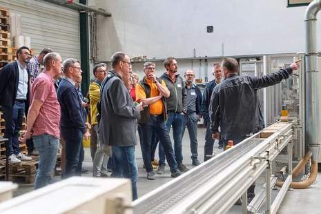 Eine Gruppe von Menschen steht in einer Werkhalle an einer Maschen. Vor der Maschine erklärt ein Mann etwas.