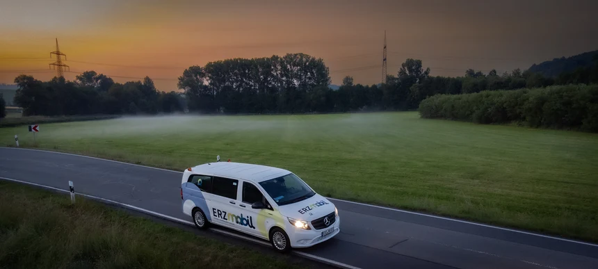 Ein weißer Kleinbus fährt auf einer Straße im ländlichen Raum. Im Hintergrund geht die Sonne unter.