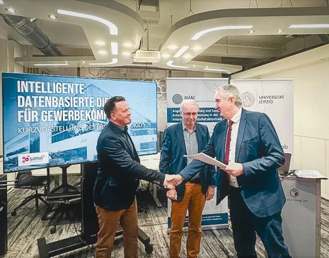 Drei Männer stehen vor einem Bildschirm und Roll-Ups. Zwei der Männer reichen sich die Hand.