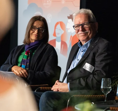 Staatssekretärin Meyer und Professor Hasinger sitzen nebeneinander und schauen ins Publikum. Im Hintergrund steht ein Banner in blau und orange.