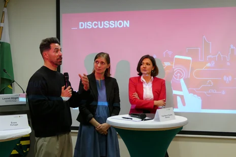 Drei Menschen stehen vor einer Leinwand an einem Tisch und diskutieren