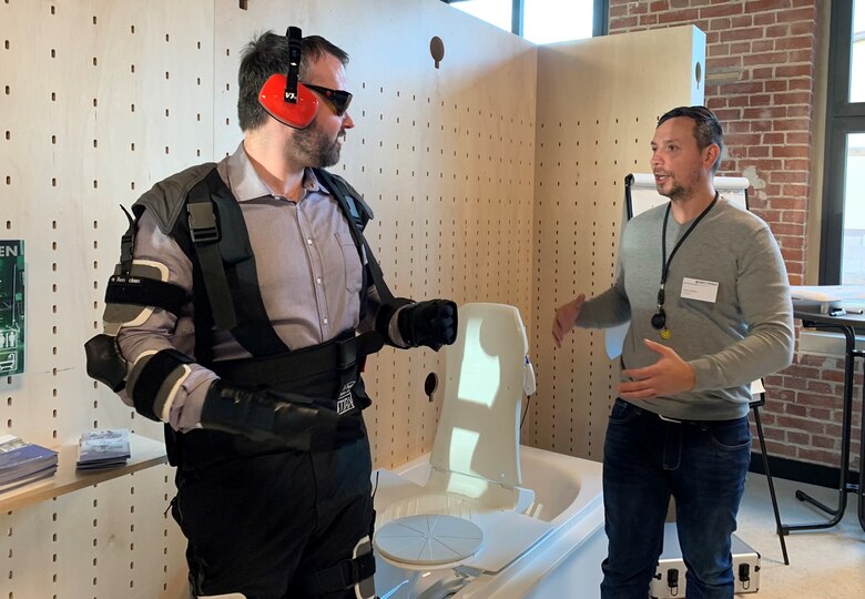 Zwei Männer stehen in einem Raum und unterhalten sich. Einer trägt eine Brille, Ohrenschätzer und verschieden Ausrüstungsgegenstände an den Armen und Beinen.