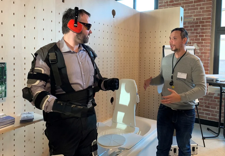 Zwei Männer stehen in einem Raum und unterhalten sich. Einer trägt eine Brille, Ohrenschätzer und verschieden Ausrüstungsgegenstände an den Armen und Beinen.