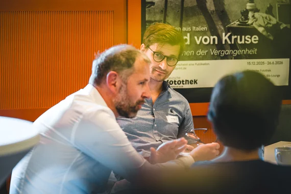 An einem Tisch sitzen zwei Männer sowie eine Frau und unterhalten sich.